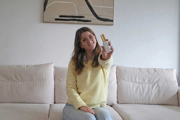 Smiling woman in yellow sweater sitting on beige sofa holding skin care serum bottle